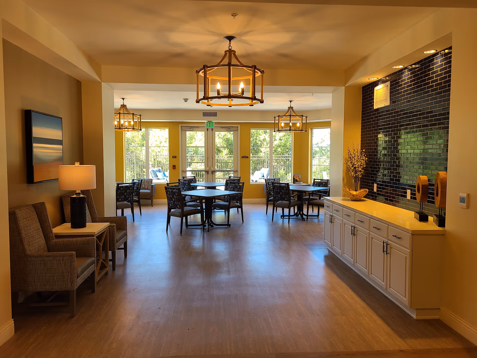 A bright and spacious dining area with several tables and chairs arranged near large windows that let in natural light. The room features warm yellow walls, modern chandeliers, a sideboard with decorative items, and comfortable seating including armchairs and a lamp on a side table.