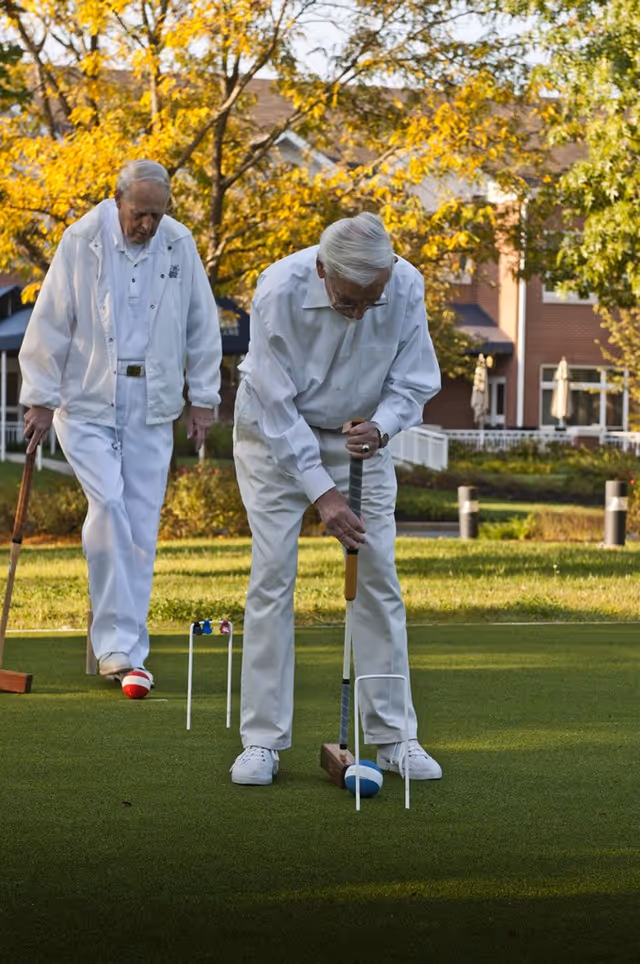 Two elderly men dressed in white playing croquet on a green lawn with a building and trees with autumn foliage in the background.