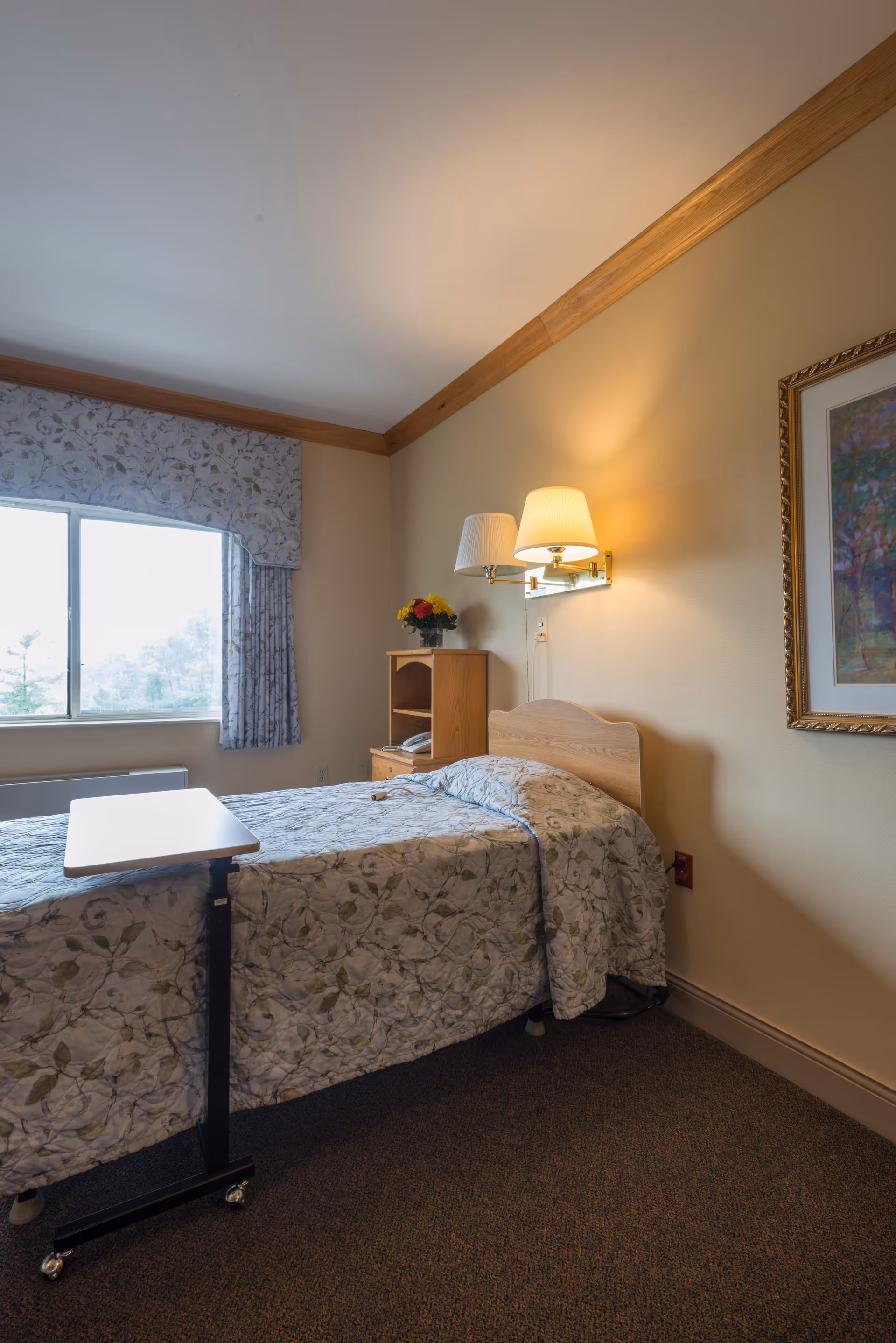 A single bed with floral patterned bedding in a room with beige walls and a window with floral curtains. There is a small wooden nightstand with a vase of flowers and a wall-mounted lamp above the bed. A framed picture hangs on the wall to the right.