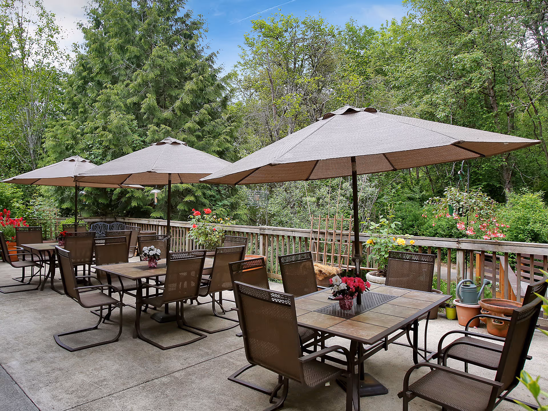 Outdoor patio area with multiple tables and chairs under large beige umbrellas, surrounded by lush green trees and various potted plants and flowers.