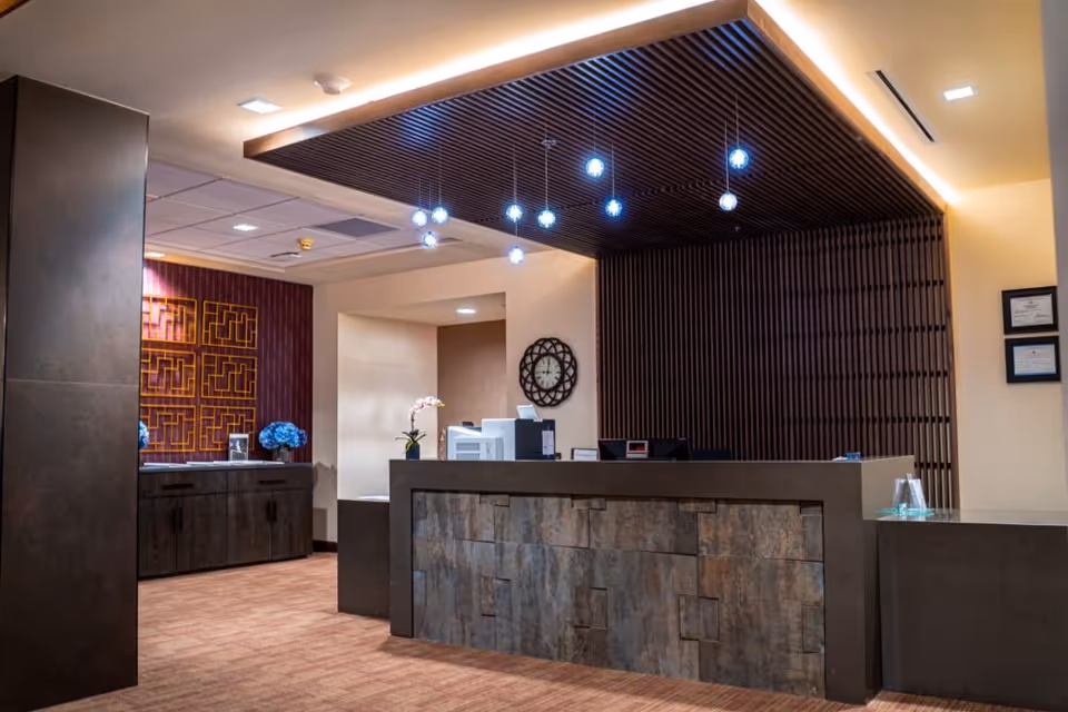 Reception area of Granite Gate Senior Living featuring a modern front desk with a textured stone facade, a decorative wall clock, hanging pendant lights, and a side cabinet with blue floral arrangements and geometric wall art.