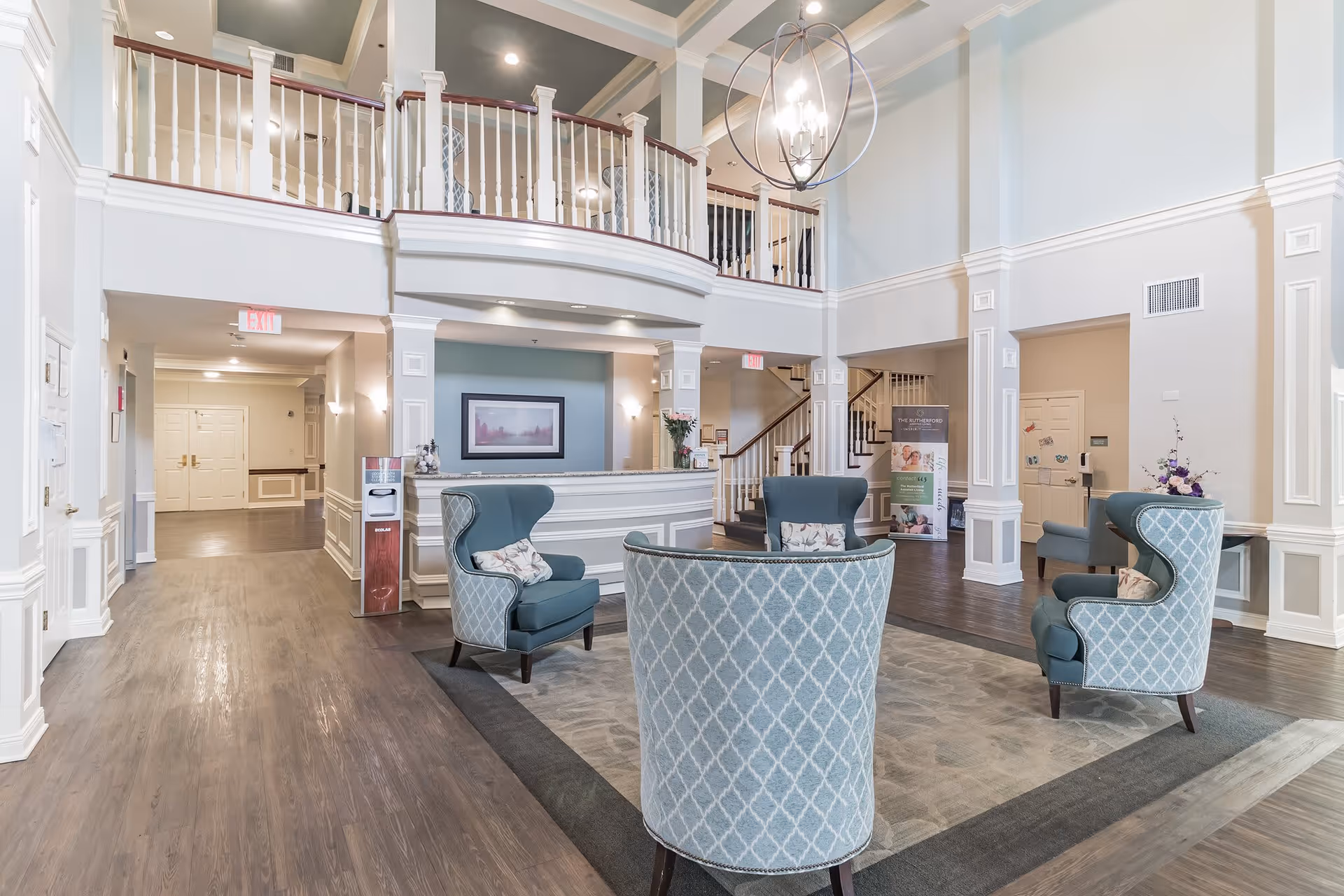 Spacious and elegant assisted living facility lobby with high ceilings, a chandelier, a reception desk, and four upholstered armchairs arranged around a rug. The area features white walls with decorative molding, wooden flooring, and a staircase leading to an upper level.