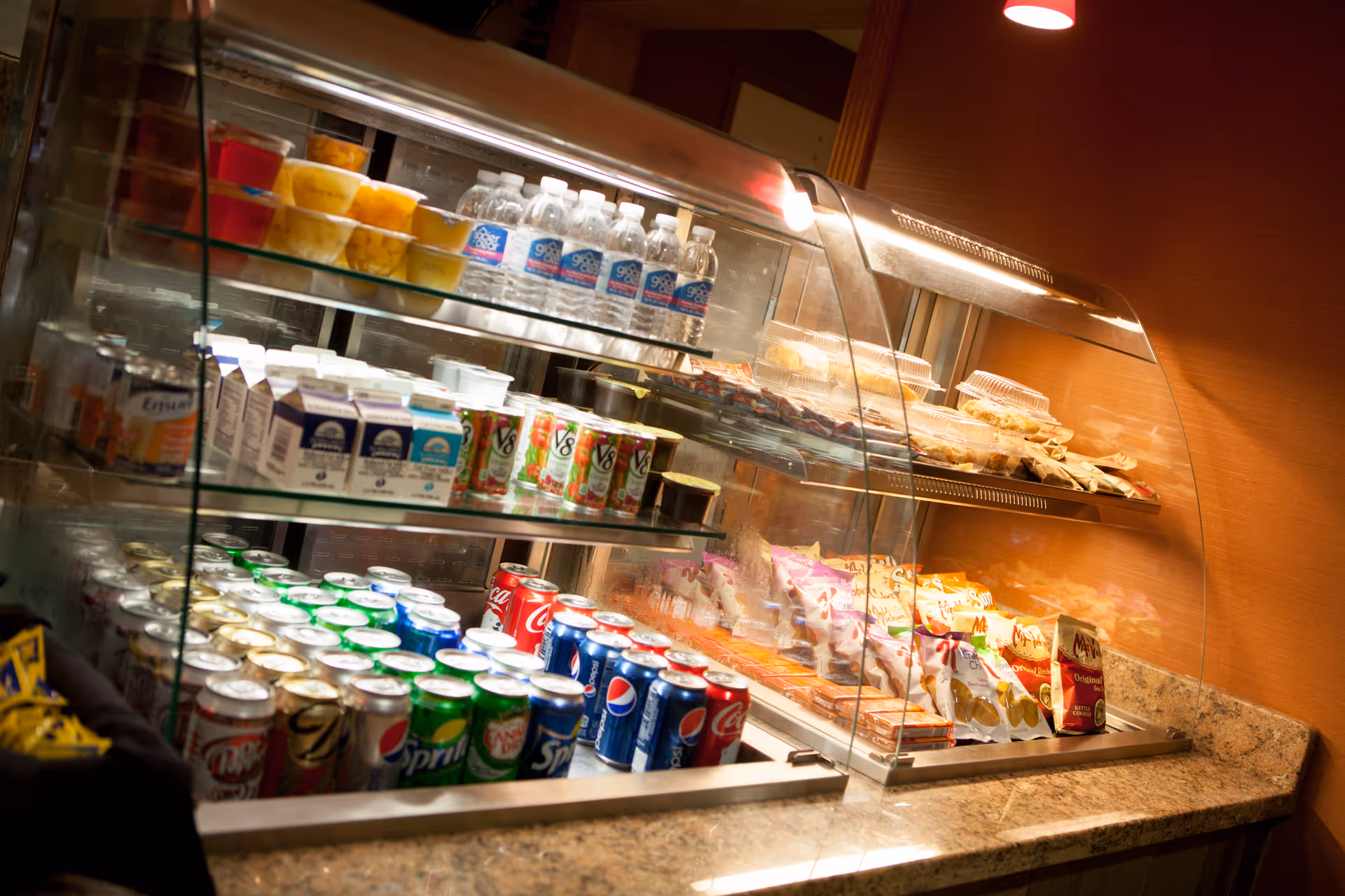A glass display case filled with various beverages and snacks including canned sodas, bottled water, fruit cups, juice cartons, and packaged chips, set on a granite countertop in a warmly lit room.