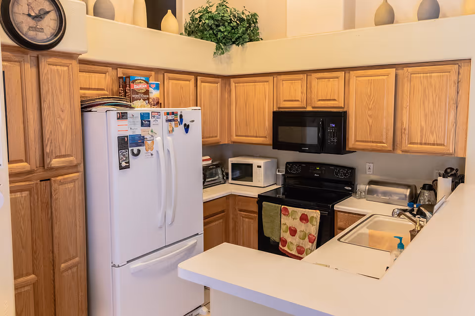 A kitchen with wooden cabinets, a white refrigerator with magnets and notes, a black microwave above a black stove with colorful towels hanging on the handle, a white countertop with a sink, a toaster oven, a microwave, and various kitchen items. There is a clock on the wall and decorative vases and greenery on a ledge above the cabinets.