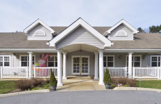 Front entrance of a single-story senior living building with a covered porch supported by white columns and a pink inflatable bunny on the left railing.