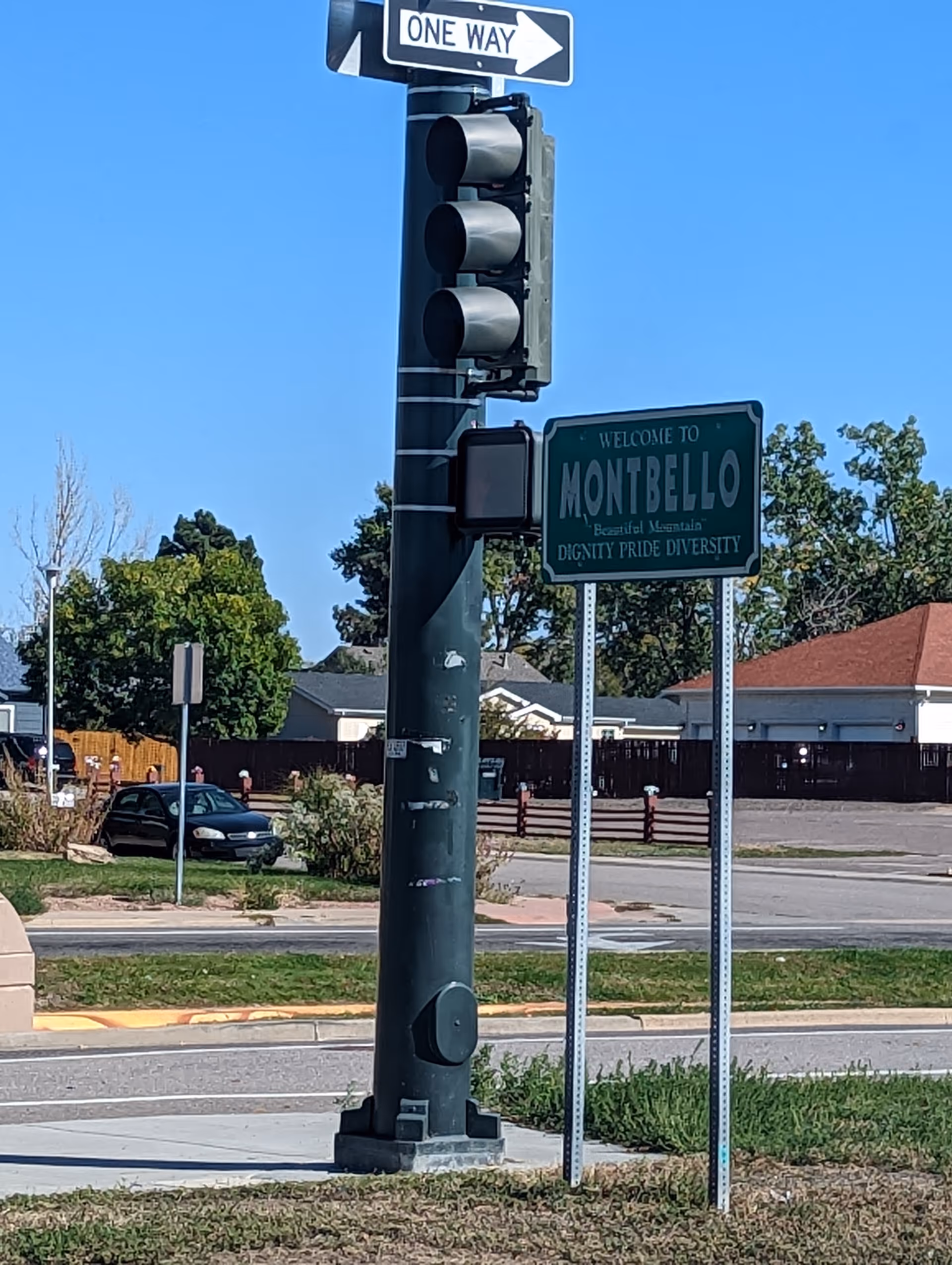 A street scene showing a traffic light pole with a one way sign pointing right and a green welcome sign that reads 'Welcome to Montbello, Beautiful Mountains, Dignity Pride Diversity'. There are trees, houses, and a parked car in the background under a clear blue sky.