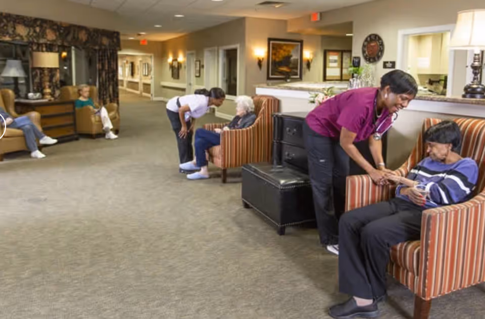 A hallway in an assisted living facility with elderly residents sitting in armchairs along the walls. Two caregivers are interacting warmly with two elderly women seated in striped armchairs near a reception desk. The area is well-lit with wall sconces and lamps, and there are framed pictures on the walls.