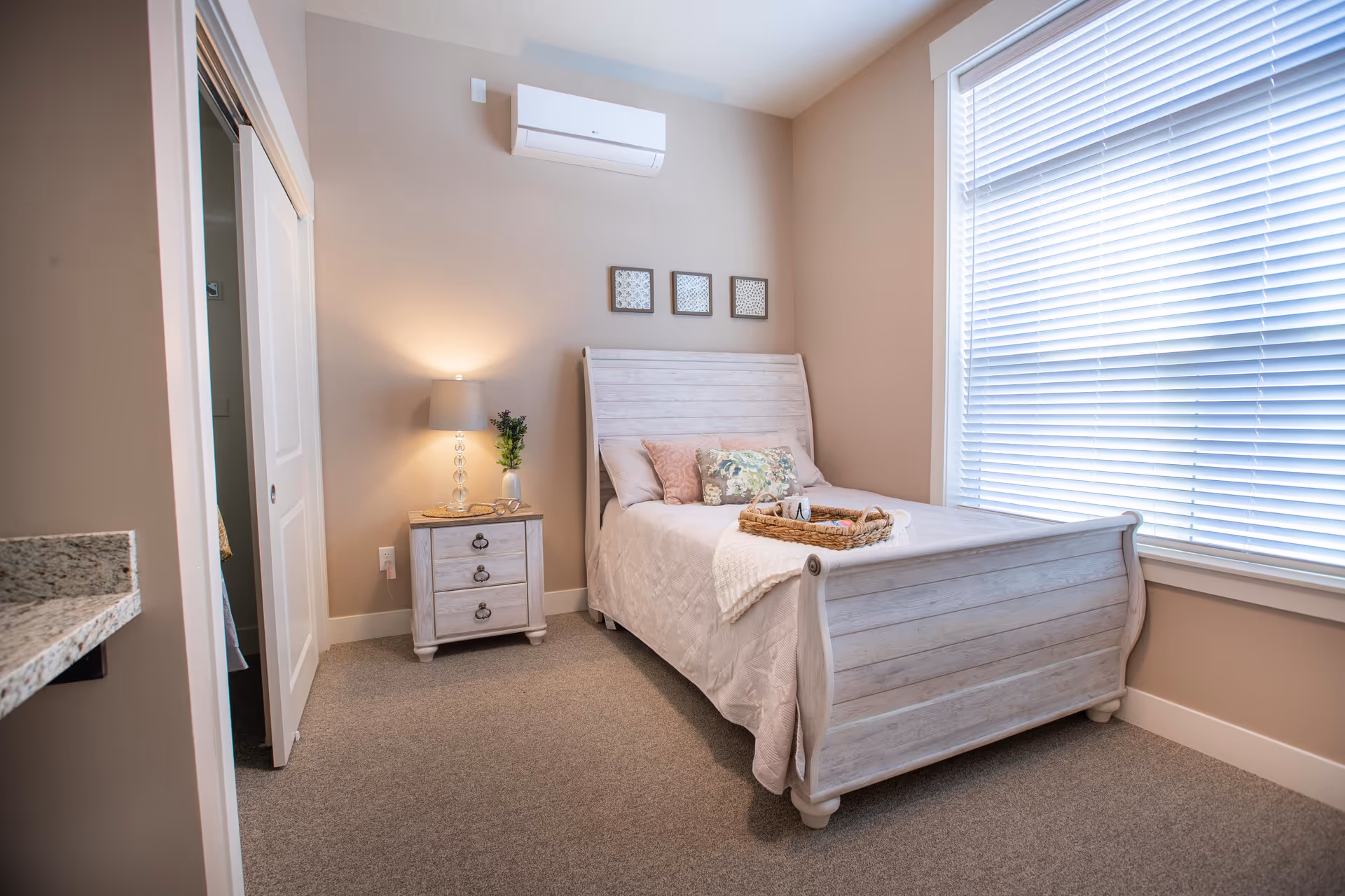 A cozy bedroom with a light wooden bed frame and matching nightstand. The bed is made with a white quilt, decorative pillows, and a wicker tray holding a mug and other items. A lamp and a small plant sit on the nightstand. The room has beige walls, a large window with white blinds, and a wall-mounted air conditioning unit above the bed.