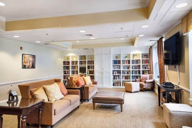 A cozy living room area with two brown sofas adorned with various cushions, a brown ottoman, and a side table with a decorative item. The back wall features built-in bookshelves filled with books and decorative pieces. There is a large flat-screen TV mounted on the right wall near a window with curtains, and a comfortable armchair with an ottoman is positioned near the bookshelves. The room has warm lighting and a neutral color palette.