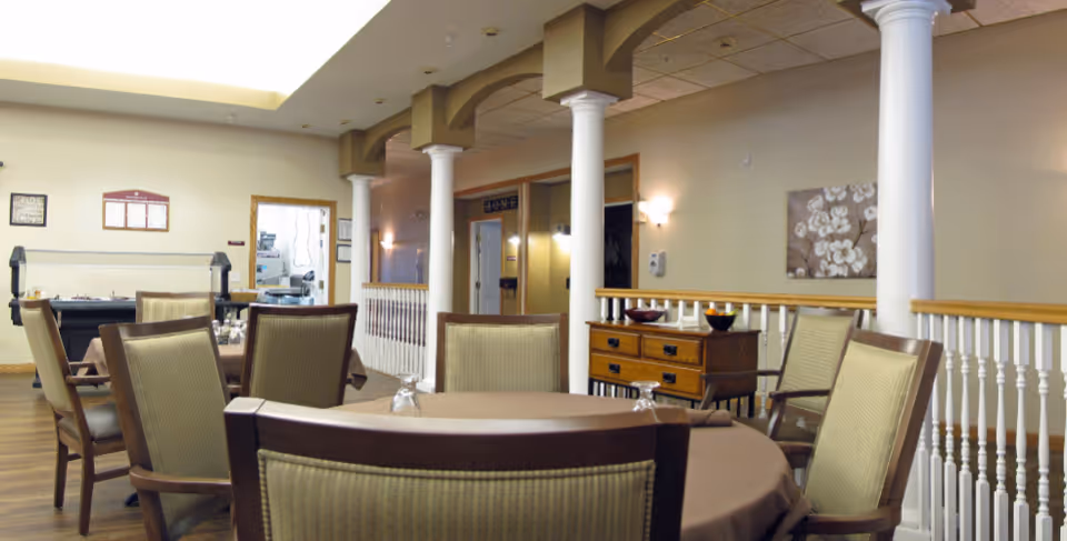 Interior dining area with round tables and upholstered chairs, decorative columns, and a buffet in a senior living facility.