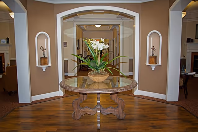 A round wooden table with a glass top holding a potted plant with white flowers is centered in a hallway with tan walls and wooden flooring. Two small wall niches with decorative sculptures are visible on either side of the hallway entrance, and a seating area with chairs and a fireplace can be seen in the background.