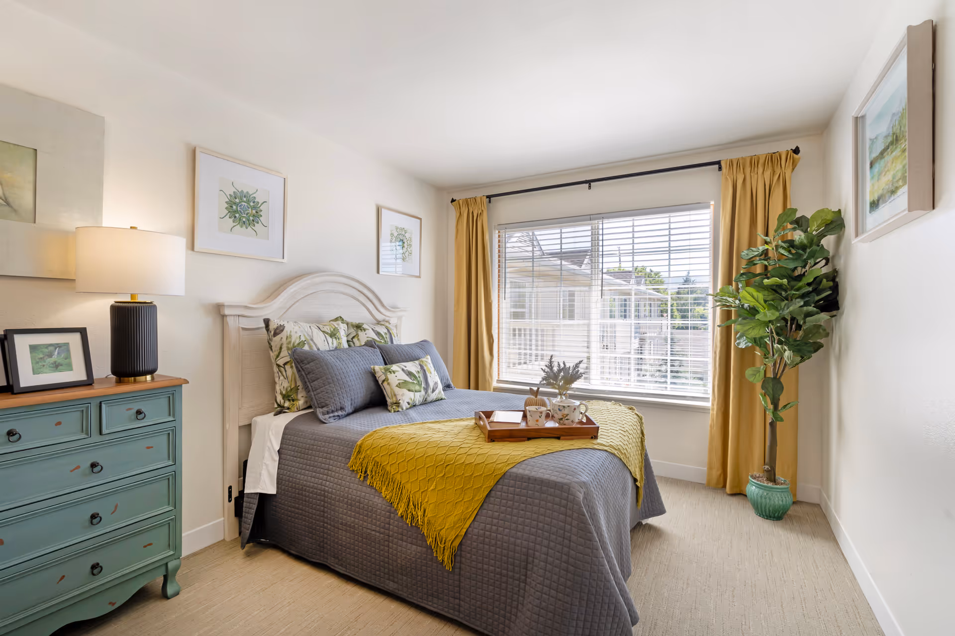 A bright and cozy bedroom with a neatly made bed featuring gray and floral pillows and a yellow throw blanket. A wooden tray with cups and a book is placed on the bed. There is a green dresser with a lamp and framed pictures on the left side, and a large window with yellow curtains letting in natural light. A potted plant is positioned in the corner near the window.