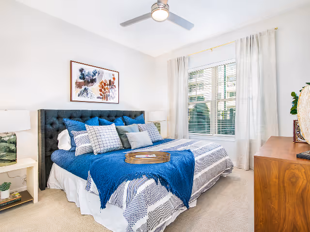A bright bedroom with a large bed featuring a tufted dark gray headboard, blue and white bedding, and multiple pillows. There is a wooden tray on the bed with a magazine. The room has beige carpet, a window with white blinds and light curtains, two bedside tables with lamps, a ceiling fan with light, a framed abstract painting above the bed, and a wooden dresser on the right side.