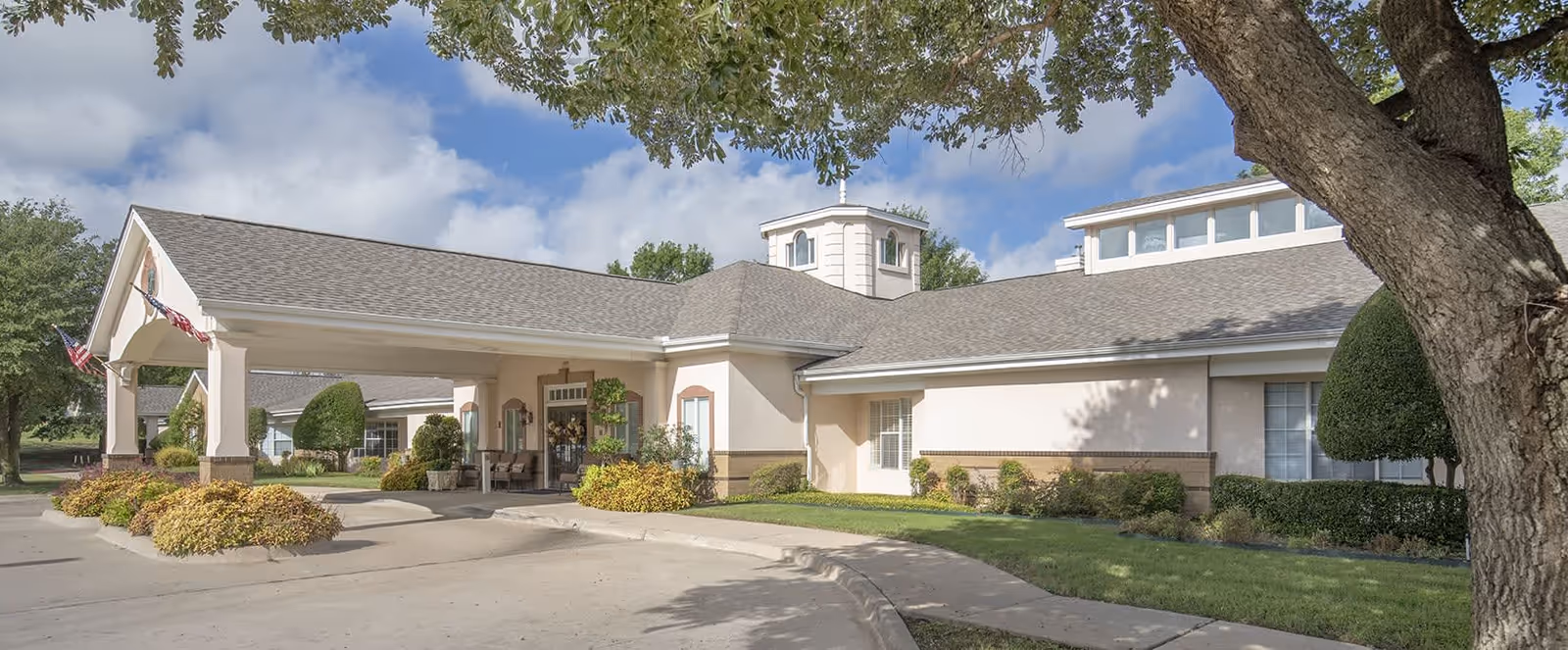 Exterior view of Brookdale Stonebridge Ranch senior living facility showing a covered entrance with two American flags, well-maintained landscaping, and a large tree in the foreground under a partly cloudy sky.