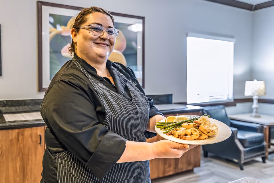 A smiling chef wearing glasses and a black apron holds a plate of food with shrimp, asparagus, rice, and lemon slices in a well-lit room with wooden cabinets, a framed picture, and a seating area with chairs and a lamp.