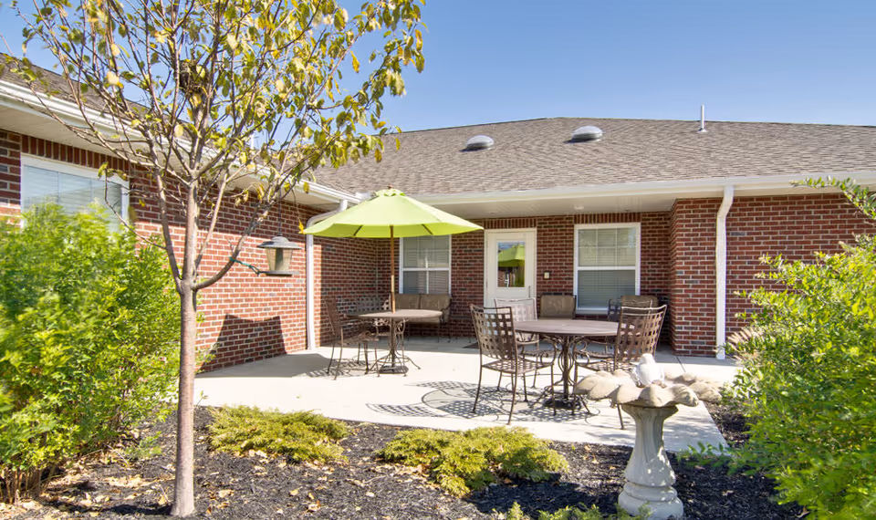 Outdoor patio area at Sugar Creek Senior Living with metal tables and chairs, one table shaded by a green umbrella, surrounded by brick building walls, windows, a door, and landscaped greenery including a small tree and bushes.