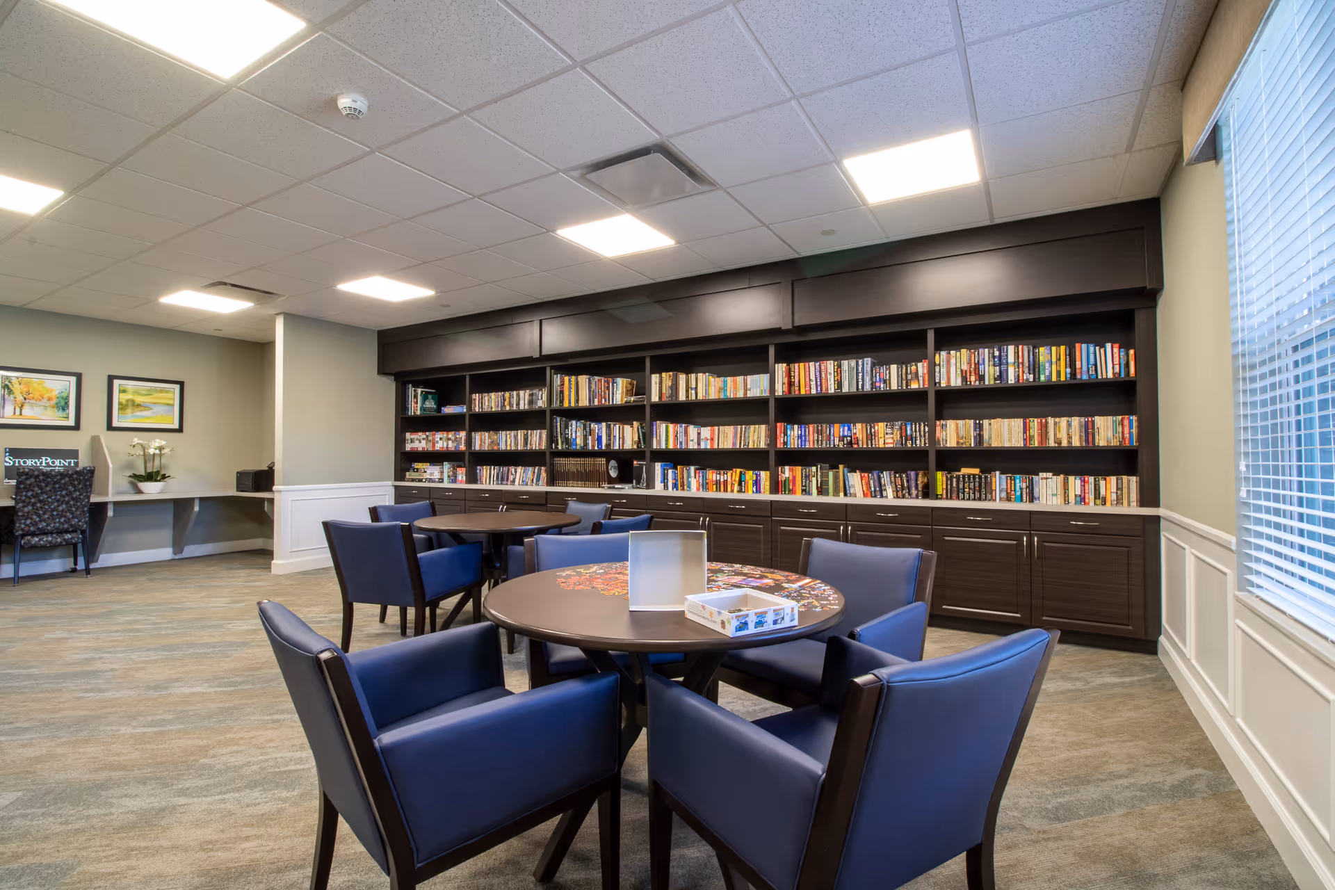 A bright common room with round tables and blue chairs in front of large bookshelves filled with books.