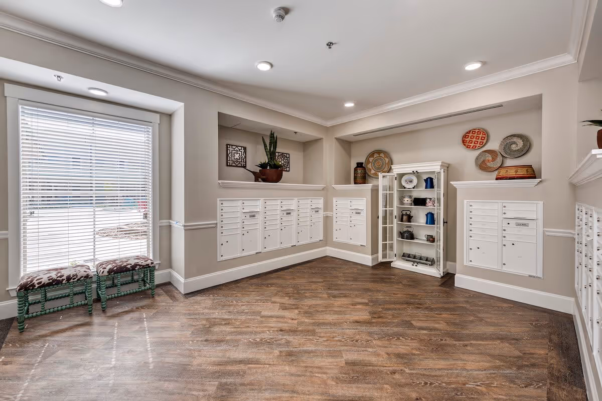 A well-lit mailroom area with multiple white mailboxes mounted on beige walls. There is a large window with white blinds on the left side, two green benches with patterned cushions below the window, and a white display cabinet with various decorative items on shelves. The floor is wooden, and the ceiling has recessed lighting.