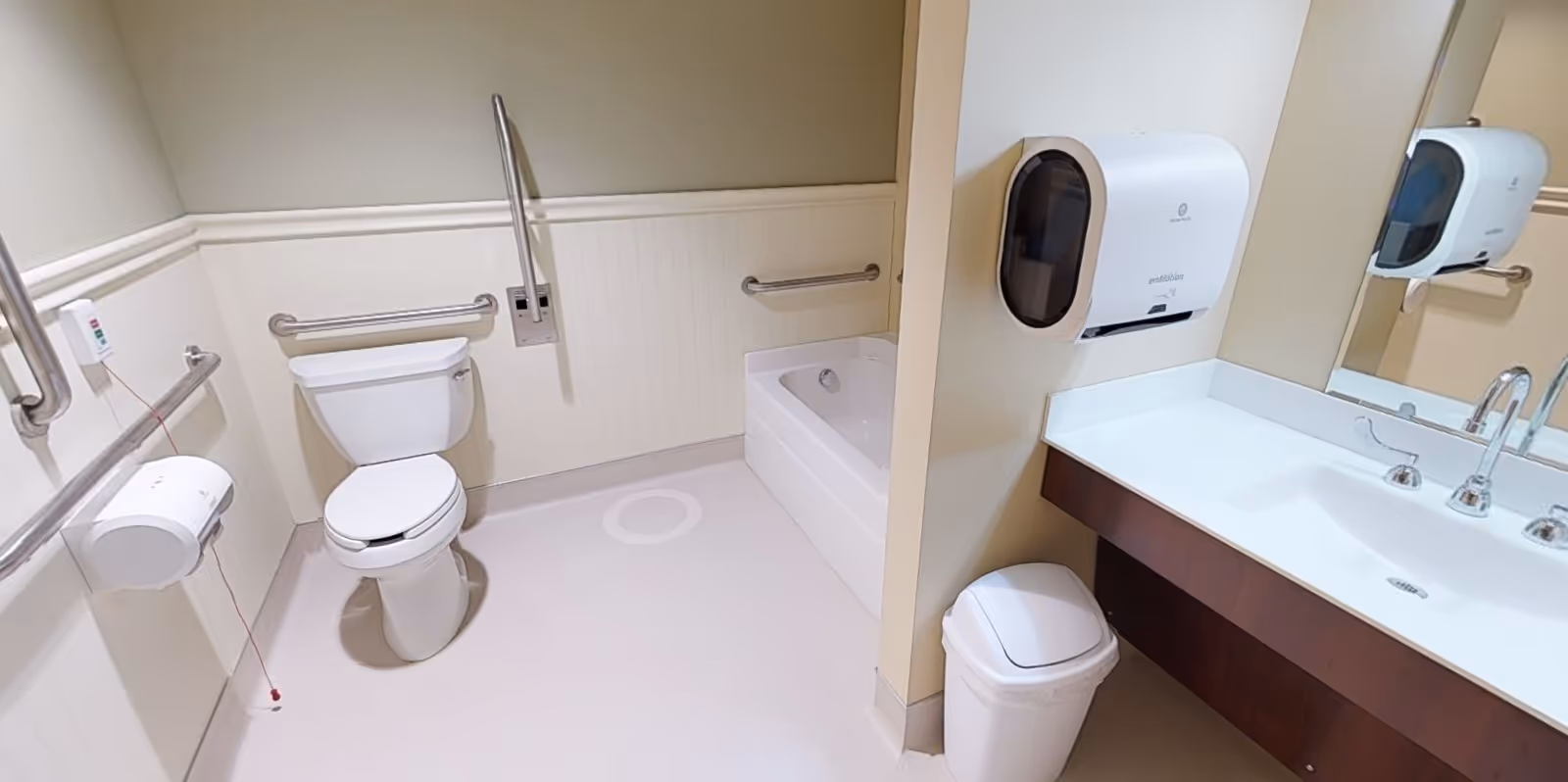 A clean and accessible bathroom featuring a toilet with grab bars on both sides, a bathtub with a grab bar, a sink with a large mirror above it, a paper towel dispenser, and a small trash bin. The walls are light-colored with wainscoting, and the floor is light-colored as well.