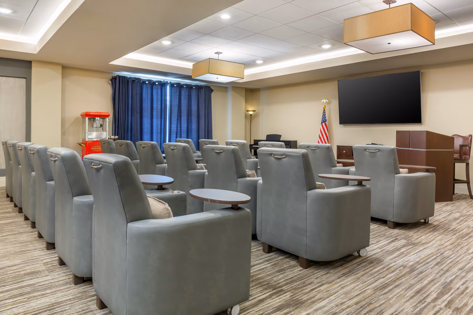 A media-style common room with rows of gray recliner chairs and small tables facing a wall-mounted TV, with a popcorn machine and an American flag.