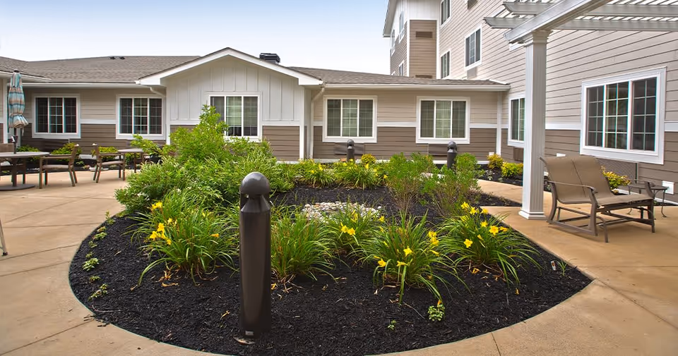 Sunny courtyard with a central planted flower bed, outdoor seating, and the surrounding senior living building.