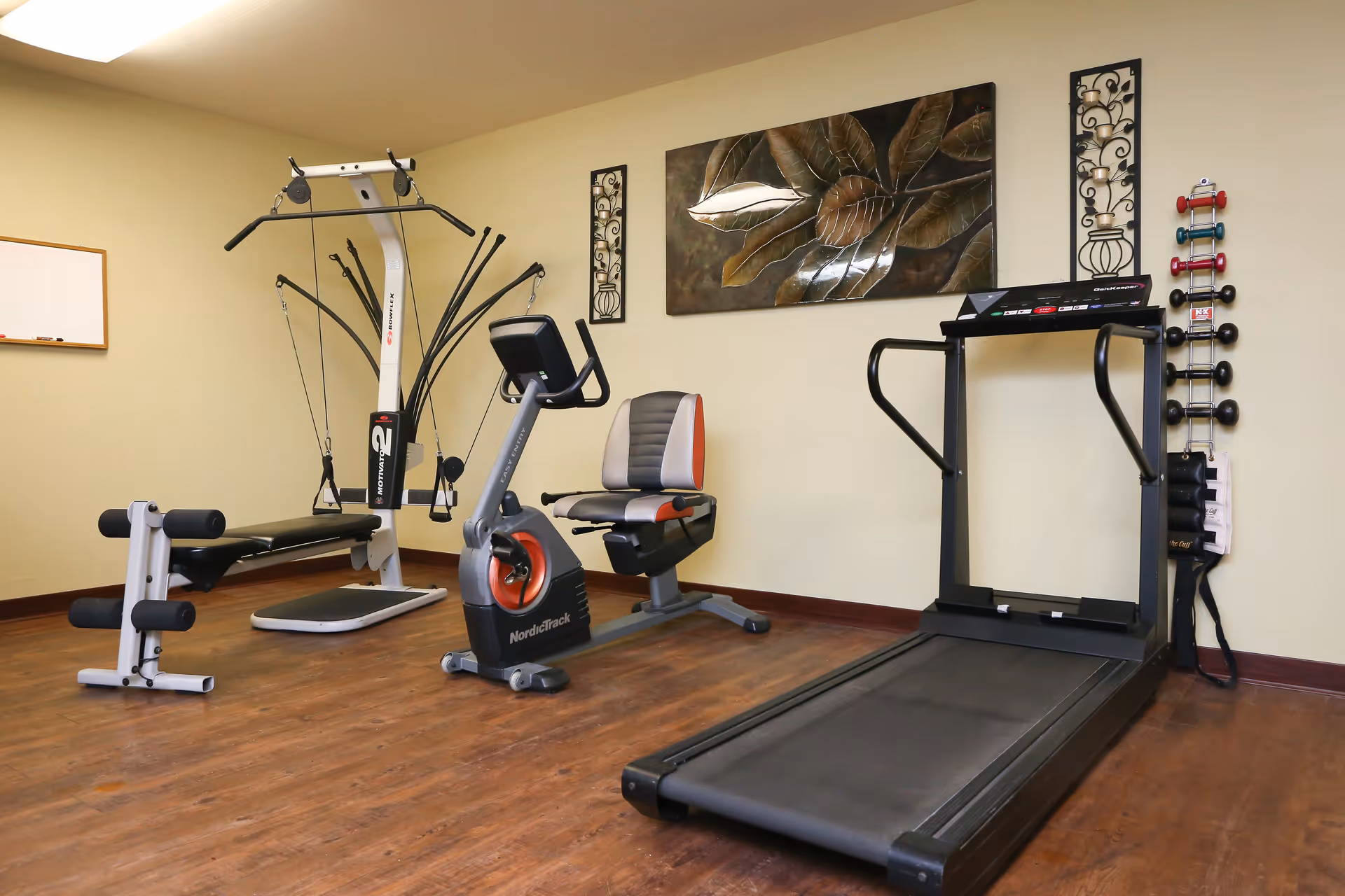 A small fitness room with exercise equipment including a treadmill, a recumbent stationary bike, a multi-function weight machine, and a bench. The room has wooden flooring, beige walls, a decorative metal wall art piece, and a rack with various dumbbells and exercise bands.