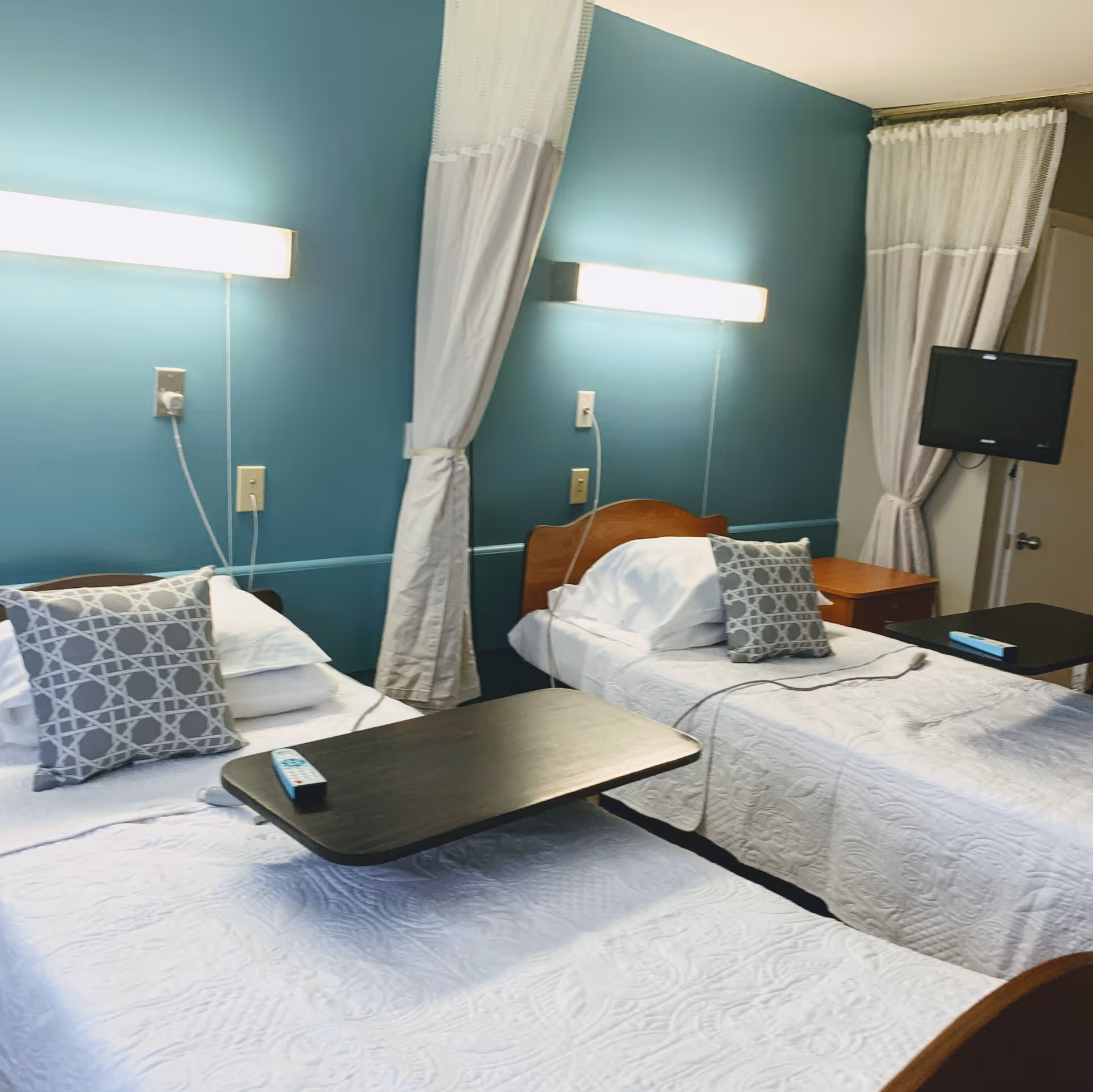 A room with two single beds, each with white bedding and a decorative gray pillow. Each bed has a dark wooden overbed table with a remote control on one. The walls are painted teal, and there are wall-mounted lights above each bed. White curtains hang between and beside the beds. A small flat-screen TV is mounted on the wall near the door.