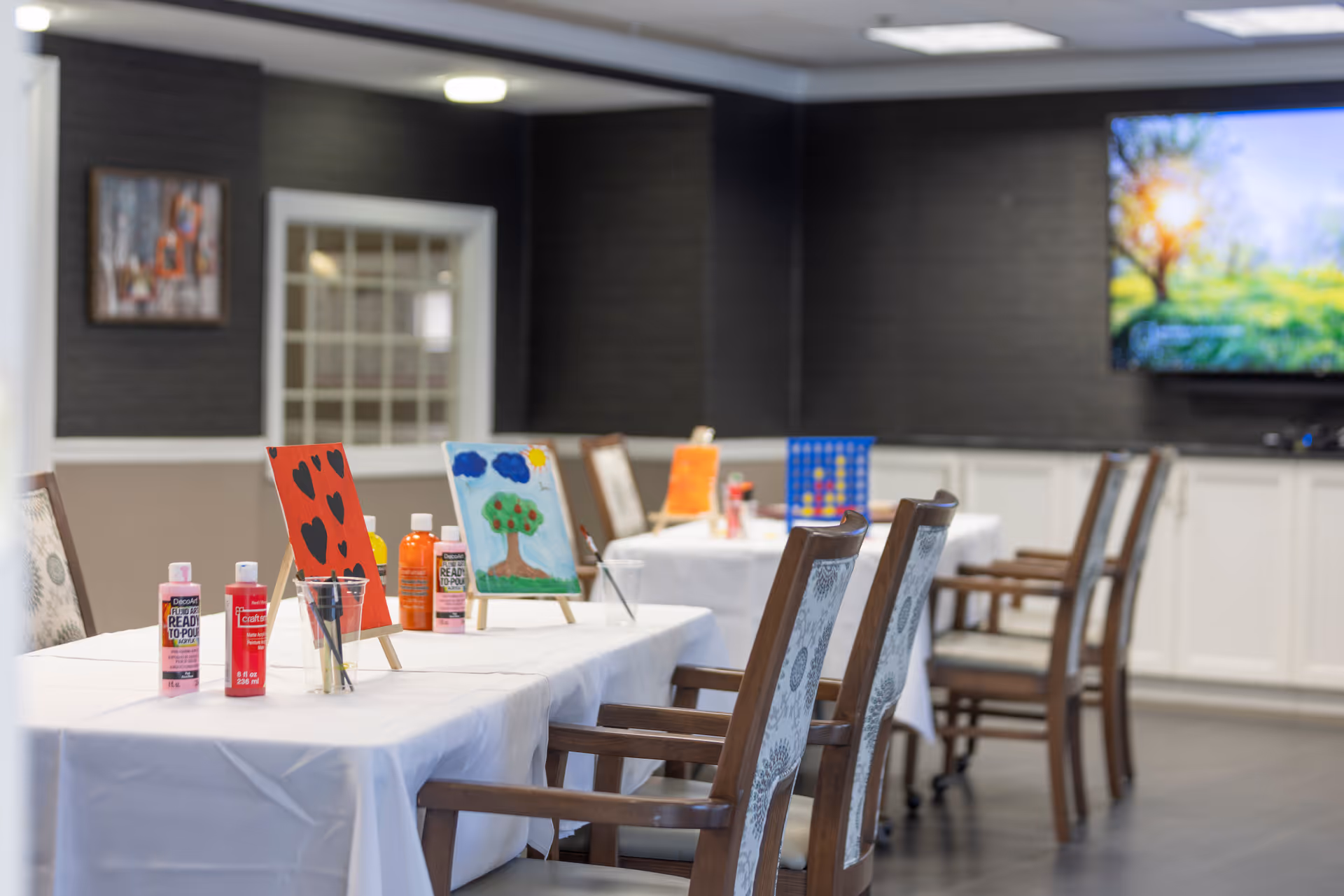 A room with tables covered in white tablecloths set up for painting activities, featuring small easels with colorful paintings, paint bottles, and brushes. The room has wooden chairs with patterned cushions, a dark accent wall, a large TV screen displaying a nature scene, and white cabinetry along the back wall.