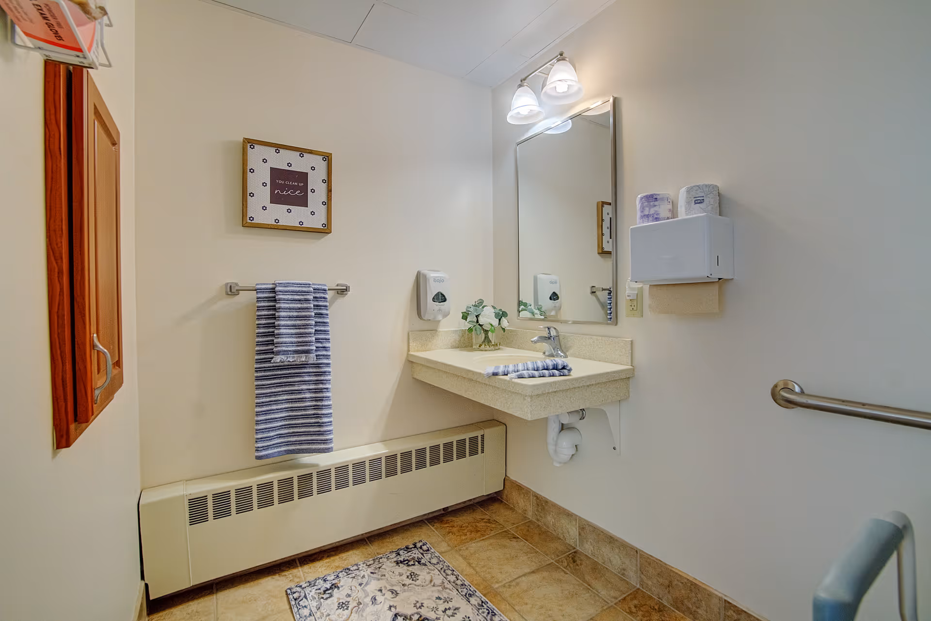 A clean and well-lit bathroom with a wall-mounted sink, a large mirror above it, a striped hand towel hanging on a towel rack, a small framed sign on the wall, a soap dispenser, a paper towel dispenser, a grab bar on the wall, and a patterned rug on the tiled floor.