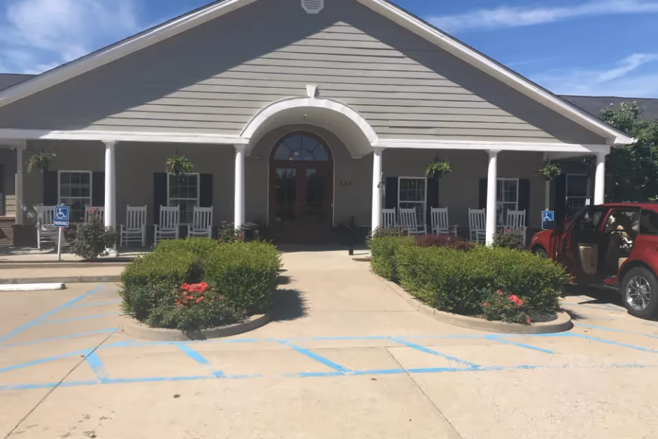 Front exterior view of Ashley Pointe Senior Living building with a covered porch featuring white rocking chairs and hanging plants. There are two handicap parking spaces in front with blue markings, and a red SUV is parked on the right side with its door open.