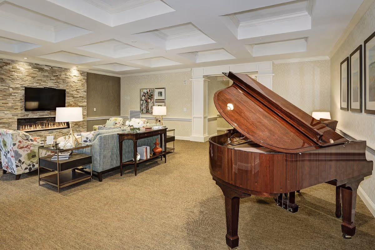 A spacious living room with a grand piano in the foreground, a seating area with floral and patterned armchairs, a sofa, side tables with lamps, a stone accent wall with a modern fireplace, and a flat-screen TV mounted above it. The room has a coffered ceiling and framed artwork on the walls.