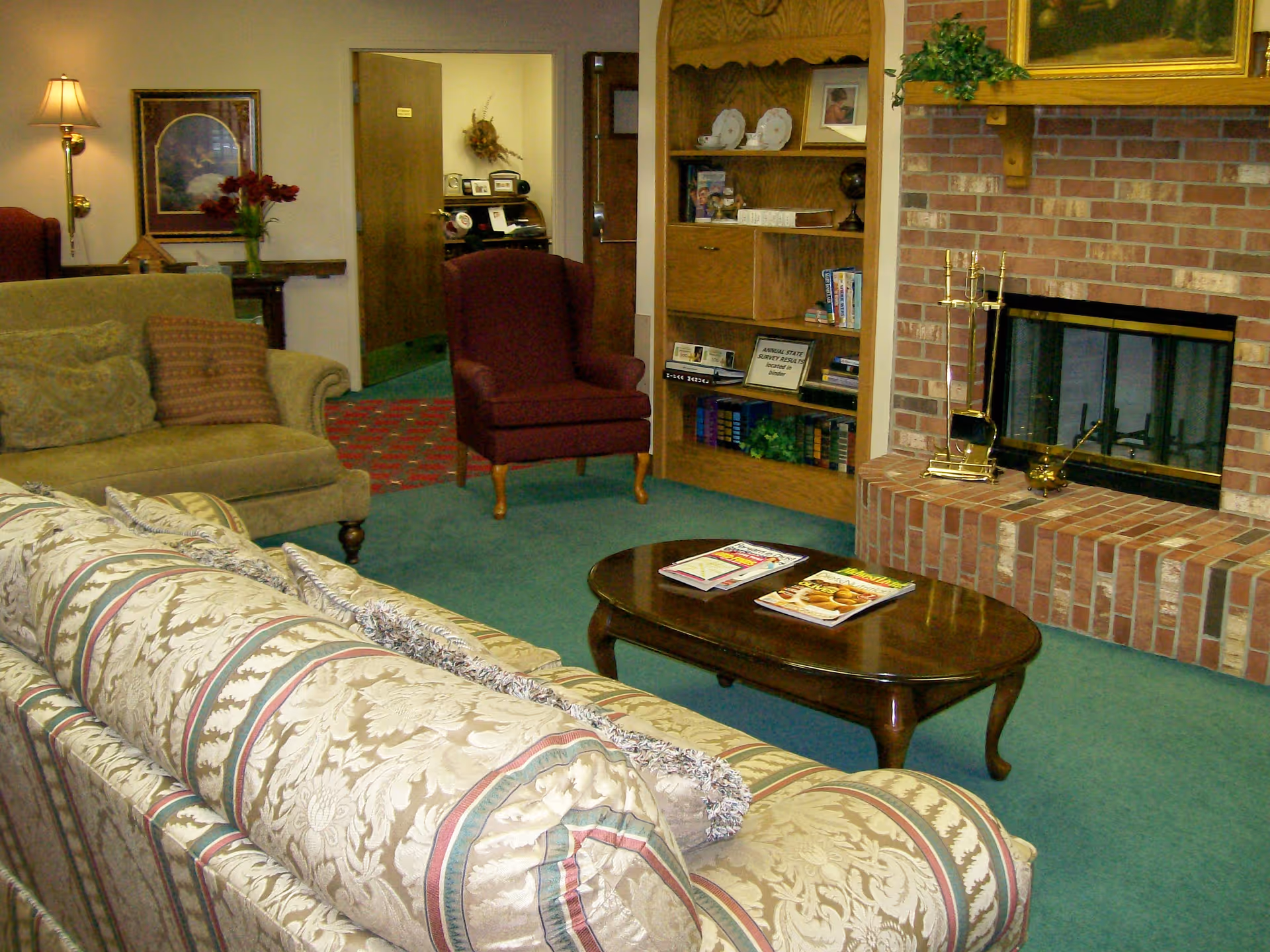 A cozy living room with a patterned sofa and a green couch, a wooden coffee table with magazines, a brick fireplace with brass fireplace tools, a wooden bookshelf filled with books and decorative items, and a burgundy armchair. The room has green carpet and walls decorated with framed pictures and a wall lamp.