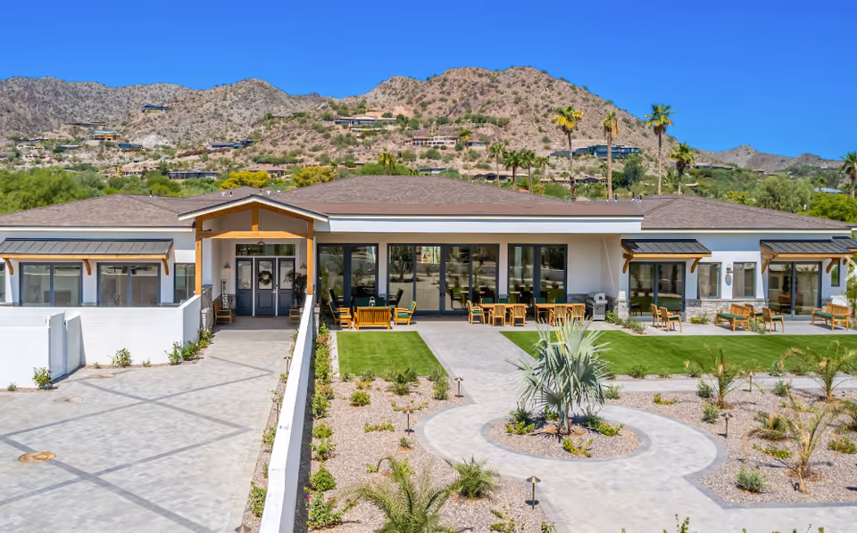 Front exterior of a single-story senior living facility with outdoor seating, landscaped pathways, and mountains in the background.