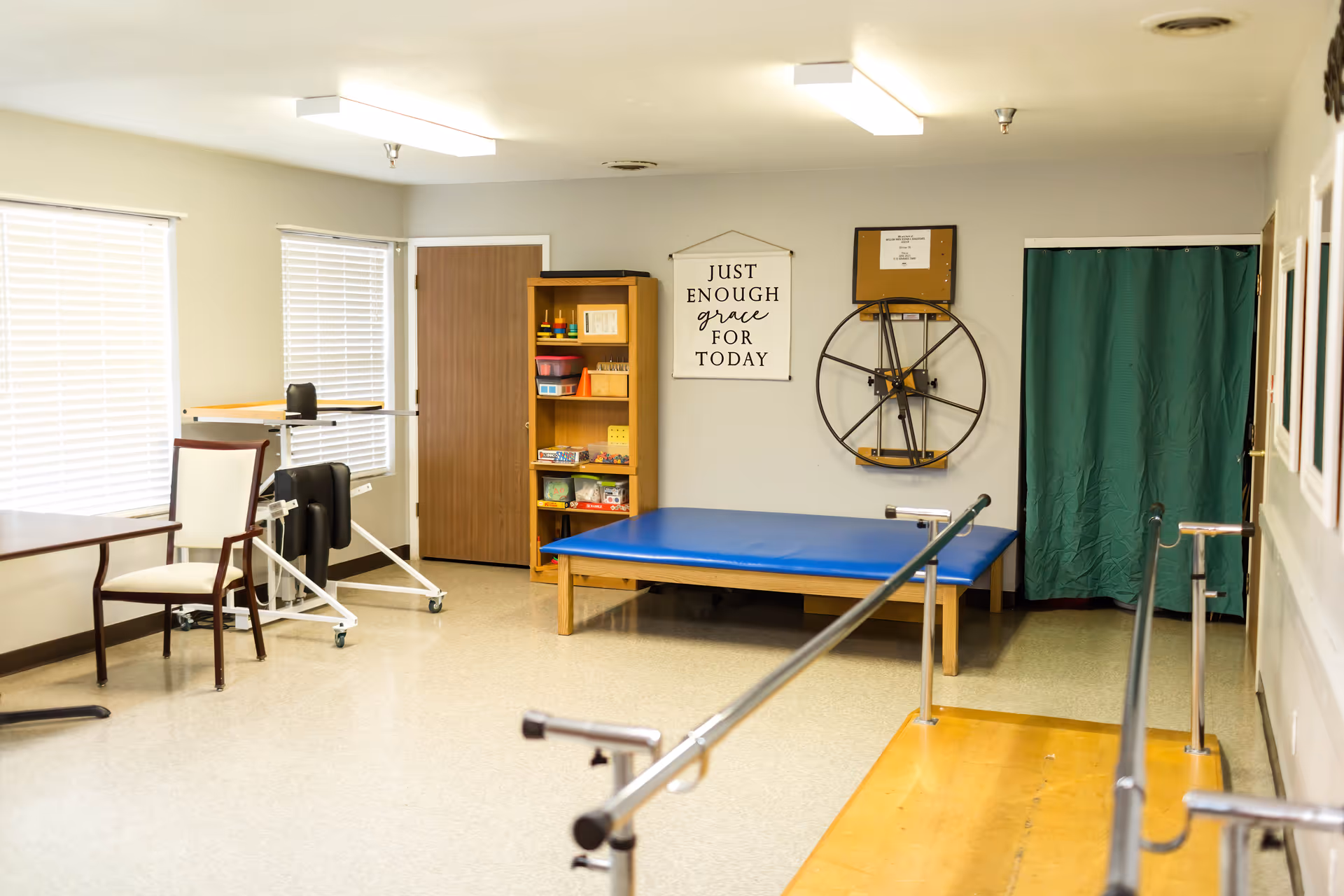 A rehabilitation therapy room with parallel bars for walking exercises, a blue padded therapy table, a wooden shelf with various therapy supplies, a chair and table near two windows with blinds, and a wall decoration that reads 'Just enough grace for today'.