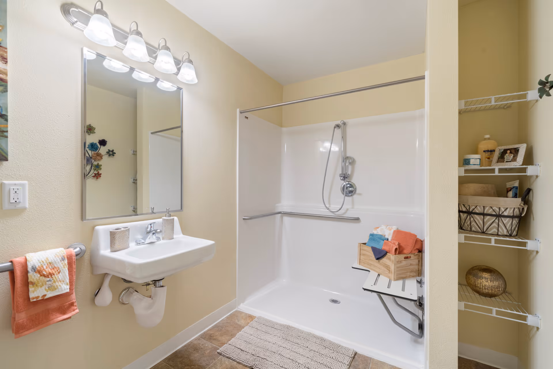 Accessible bathroom with a wall-mounted sink, mirror and vanity lights, walk-in shower with a fold-down seat and grab bar, and shelving with towels and toiletries.