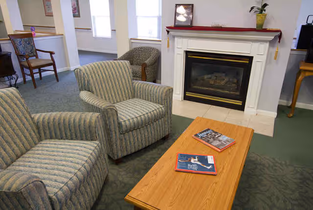 A cozy living room area with two striped armchairs and a wooden coffee table with magazines on it. There is a white fireplace with a framed picture and a potted plant on the mantel. In the background, there is another chair and windows letting in natural light.