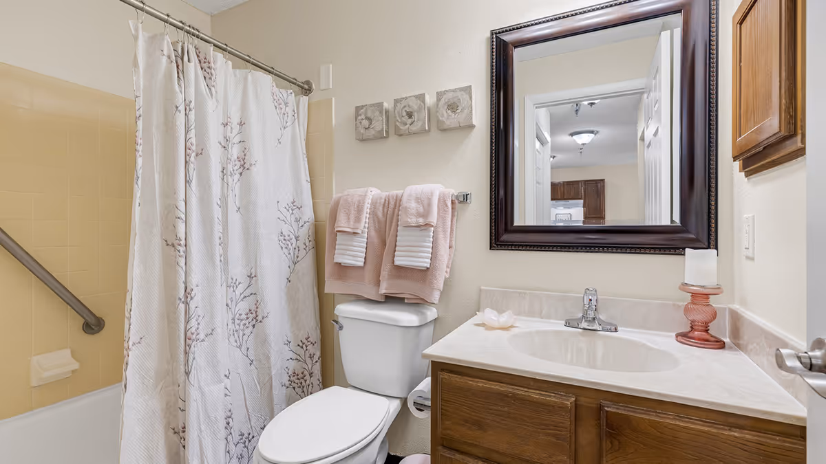 A bathroom with a white toilet, a sink with a marble countertop, a large dark-framed mirror above the sink, and a bathtub with a floral shower curtain. There are two pink towels hanging on a towel rack above the toilet, three small floral wall decorations, and a pink candle holder with a white candle on the countertop.