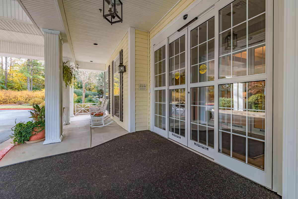 Covered entrance porch with rocking chairs, potted plants, white columns and large glass double doors.
