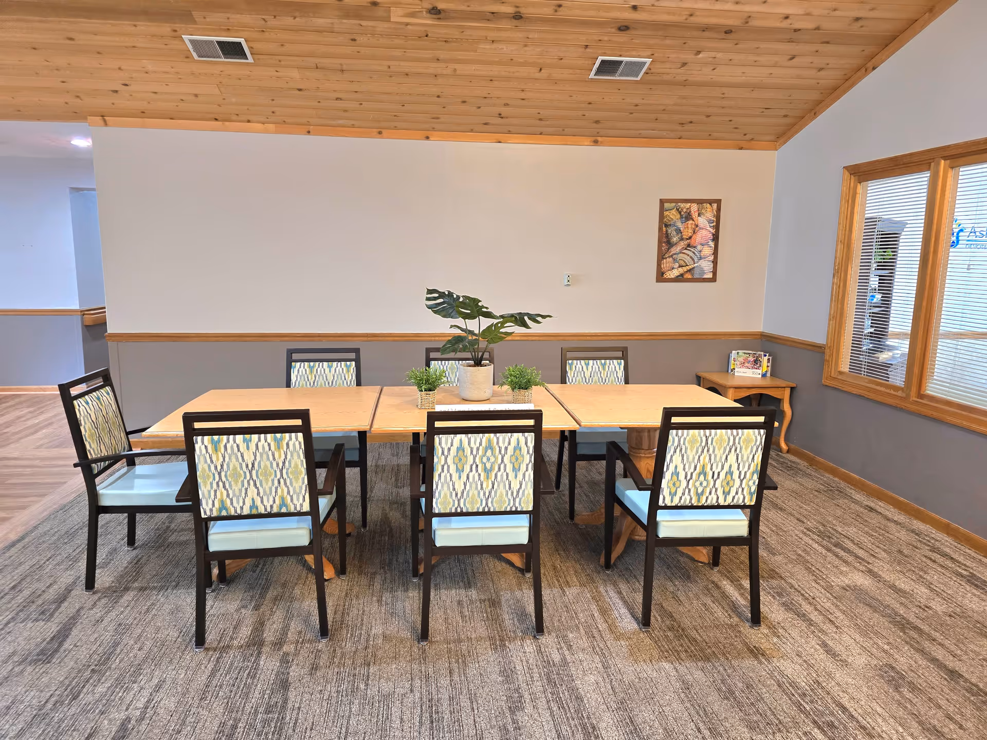A dining area with six chairs around three rectangular tables pushed together. The chairs have patterned backs and light blue seats. On the tables are three small potted plants, including one larger plant in the center. The room has a wooden ceiling, gray and white walls, carpeted floor, and a window with blinds on the right side. A small side table with magazines is in the corner.
