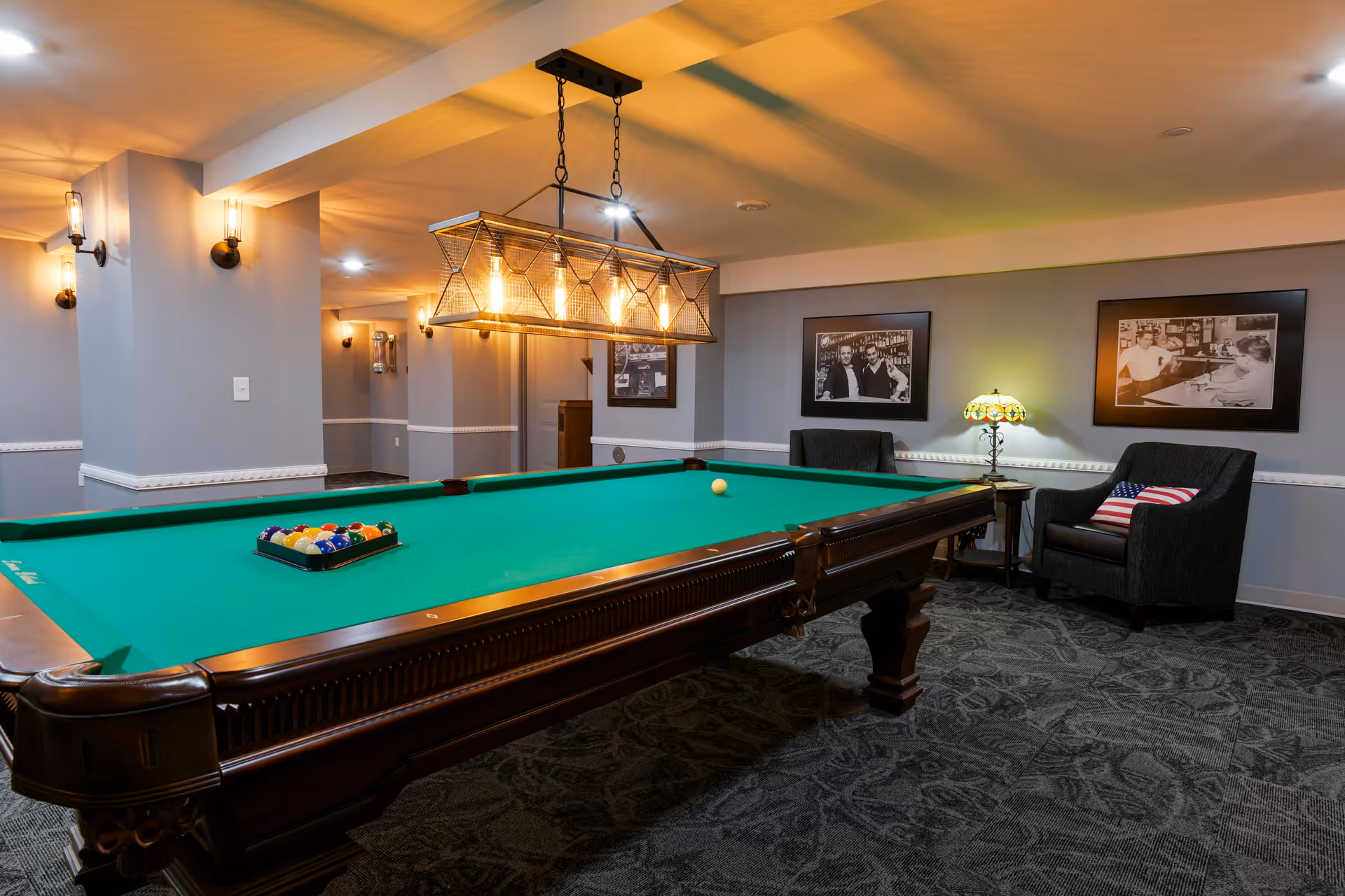 A well-lit recreational room featuring a large pool table with a green felt surface and a set of billiard balls arranged in a triangle. Above the pool table hangs a rectangular light fixture with multiple exposed bulbs. The room has gray walls adorned with black and white framed photographs. Two dark upholstered armchairs, one with an American flag pillow, sit beside a small table with a decorative lamp. The carpet has a dark patterned design, and the space has a cozy, inviting atmosphere.