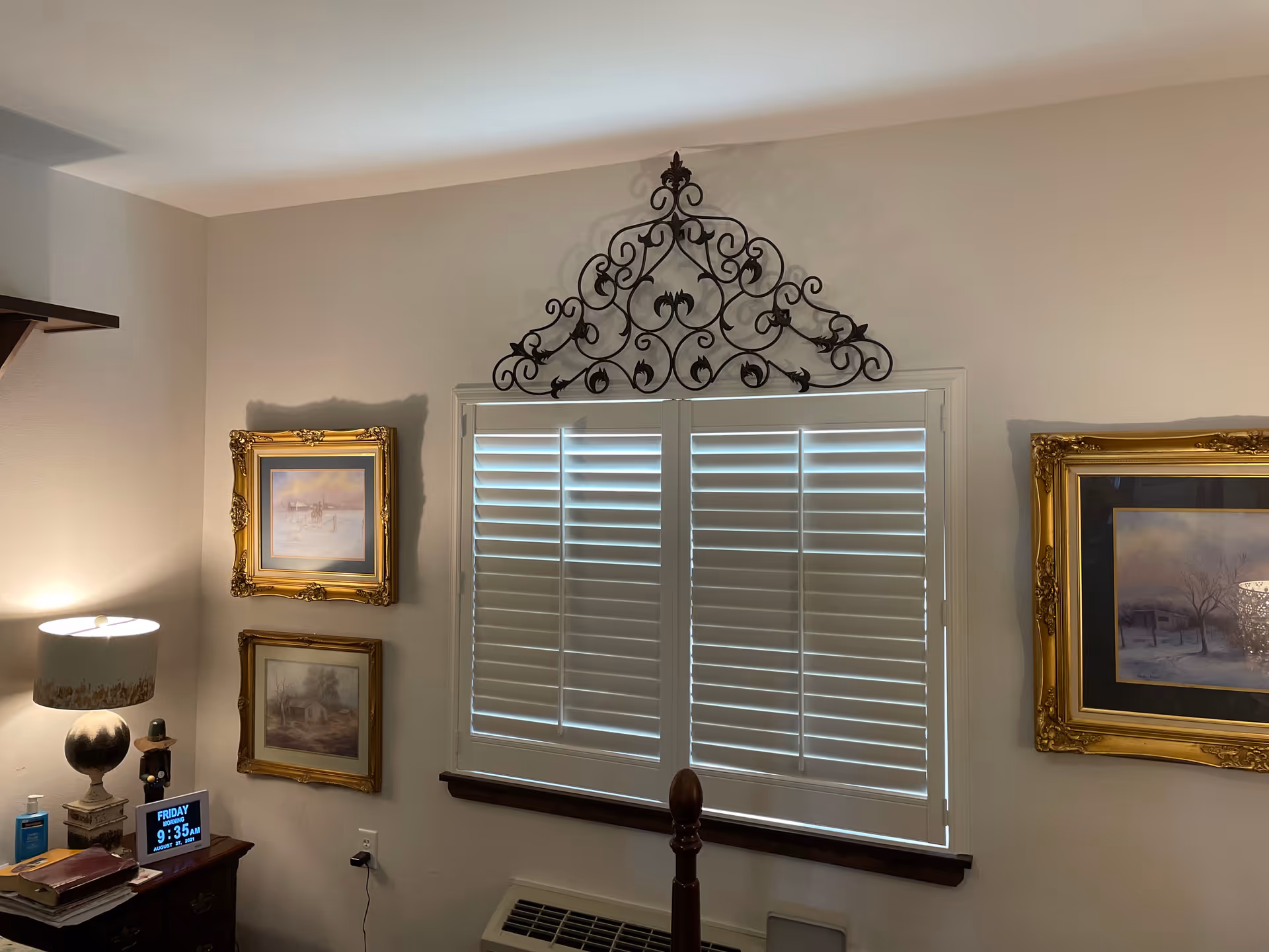 A cozy interior corner of a room featuring a window with white plantation shutters and an ornate black metal wall decoration above it. On either side of the window are framed paintings with gold-colored frames. To the left, there is a wooden nightstand with a lamp, books, and a digital clock displaying the time and date. The walls are painted light beige.