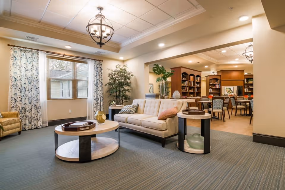 A well-lit living room area in a senior living facility featuring a beige sofa with decorative pillows, a round coffee table with a tray and vase, a side table with decorative items, a large window with floral curtains, a green armchair, and a large potted plant. In the background, there is a dining area with tables, chairs, and bookshelves filled with books and decorations.