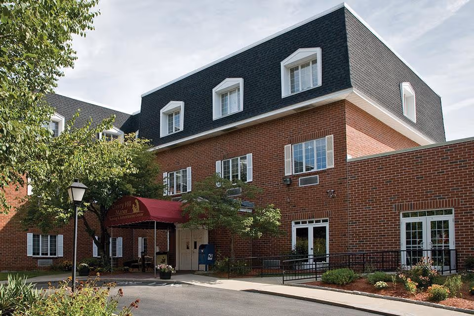 Exterior view of Cherry Hill Manor Nursing and Rehab Center, a multi-story brick building with white-trimmed windows and a black mansard roof. The entrance features a red canopy with the facility's name, surrounded by trees, shrubs, and a lamp post along a paved driveway.