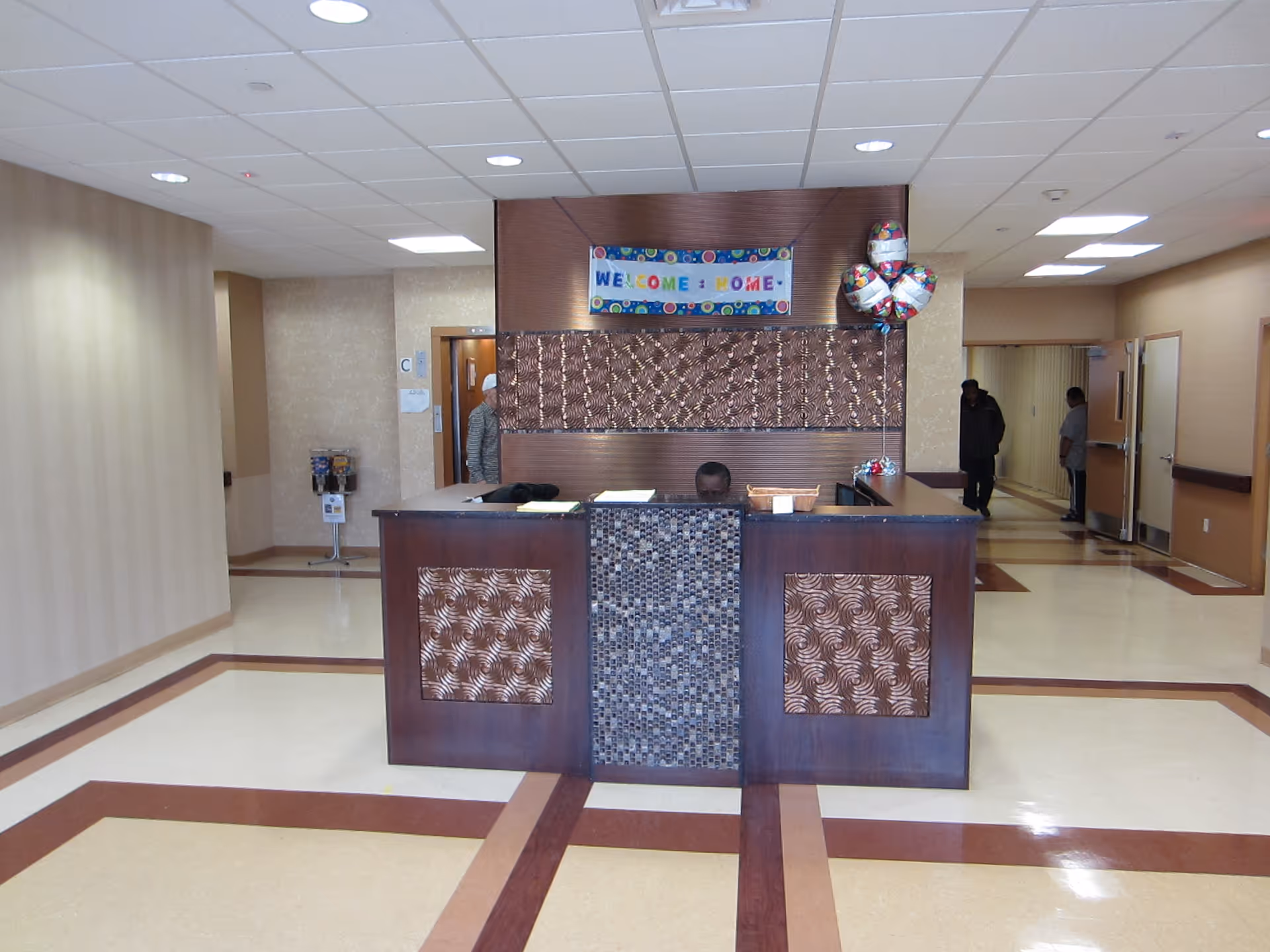 Reception area with a dark wood front desk featuring decorative panels and a mosaic center. Behind the desk is a wall with a 'Welcome Home' banner and three balloons. The floor has a beige and brown pattern, and there are people visible in the background hallway.