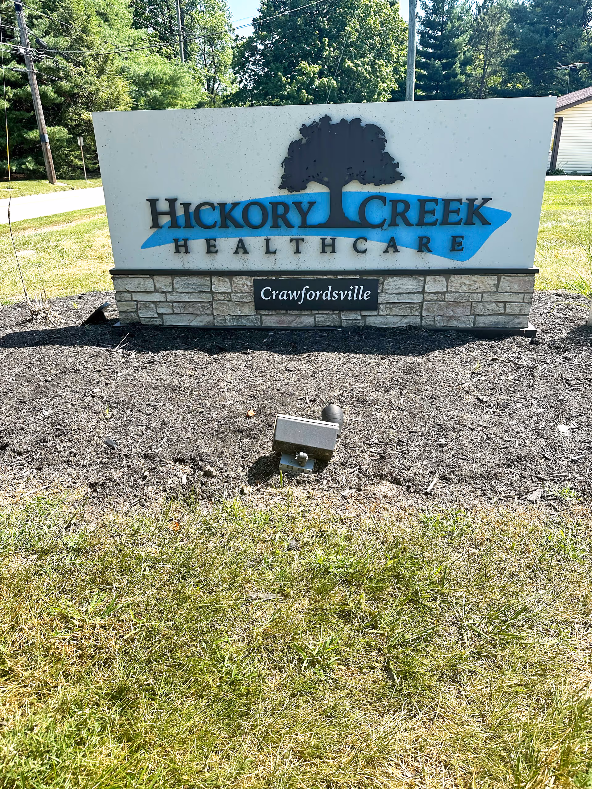 Outdoor sign for Hickory Creek Healthcare in Crawfordsville, featuring a large tree logo above the facility name on a stone and concrete base, surrounded by grass and mulch with trees and a road in the background.