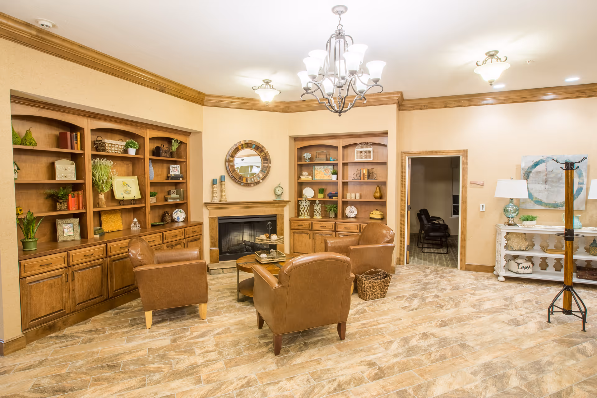 A cozy living room area with three brown leather armchairs arranged around a round wooden coffee table. Behind the chairs is a fireplace with a round decorative mirror above it, flanked by built-in wooden shelves filled with various decorative items and plants. The room has warm beige walls, a tiled floor, and a chandelier hanging from the ceiling. To the right, there is a white console table with lamps and artwork on the wall, and a wooden coat rack stands nearby.
