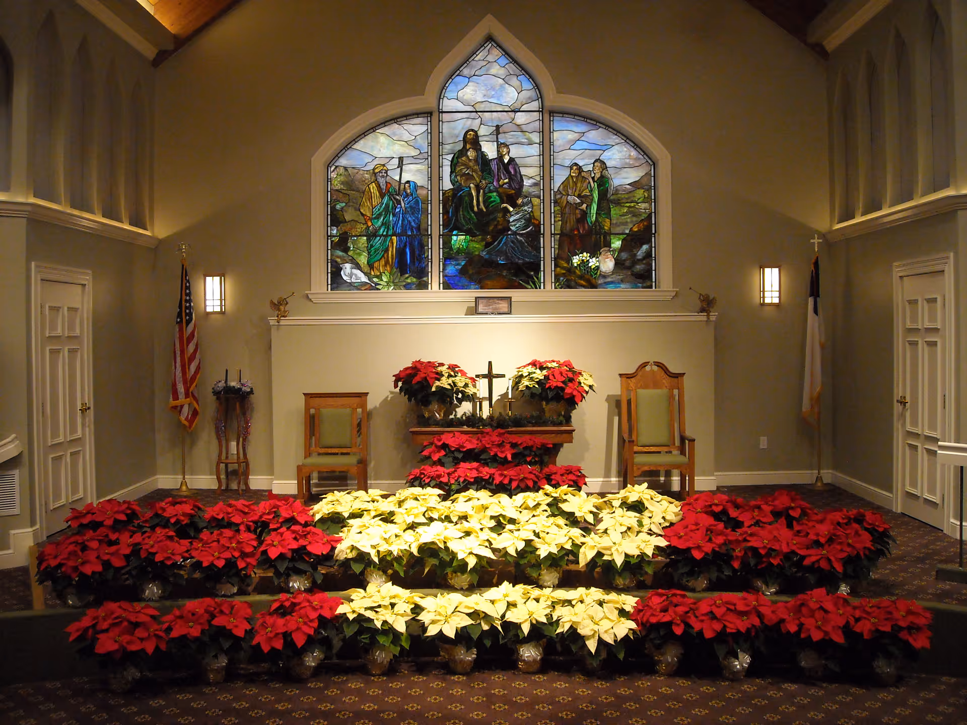 Interior view of a chapel with a stained glass window depicting religious figures above an altar adorned with red and white poinsettias. Two chairs flank the altar, and American and Christian flags stand on either side. The room has soft lighting and a carpeted floor.