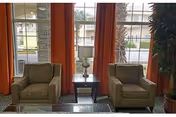 Two beige armchairs separated by a small dark wooden side table with a lamp on it, positioned in front of large windows with orange curtains. Outside the windows, parts of a building and some greenery are visible.