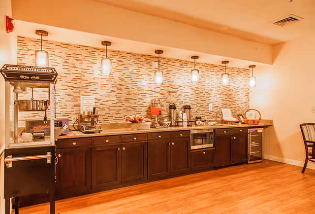 A kitchenette area with dark wood cabinets and a countertop featuring a popcorn machine, coffee dispensers, a microwave, a small refrigerator, and various snacks and supplies. The backsplash is made of small rectangular tiles in neutral tones, and five pendant lights hang from the ceiling above the counter. The floor is wooden, and a chair is partially visible on the right side.