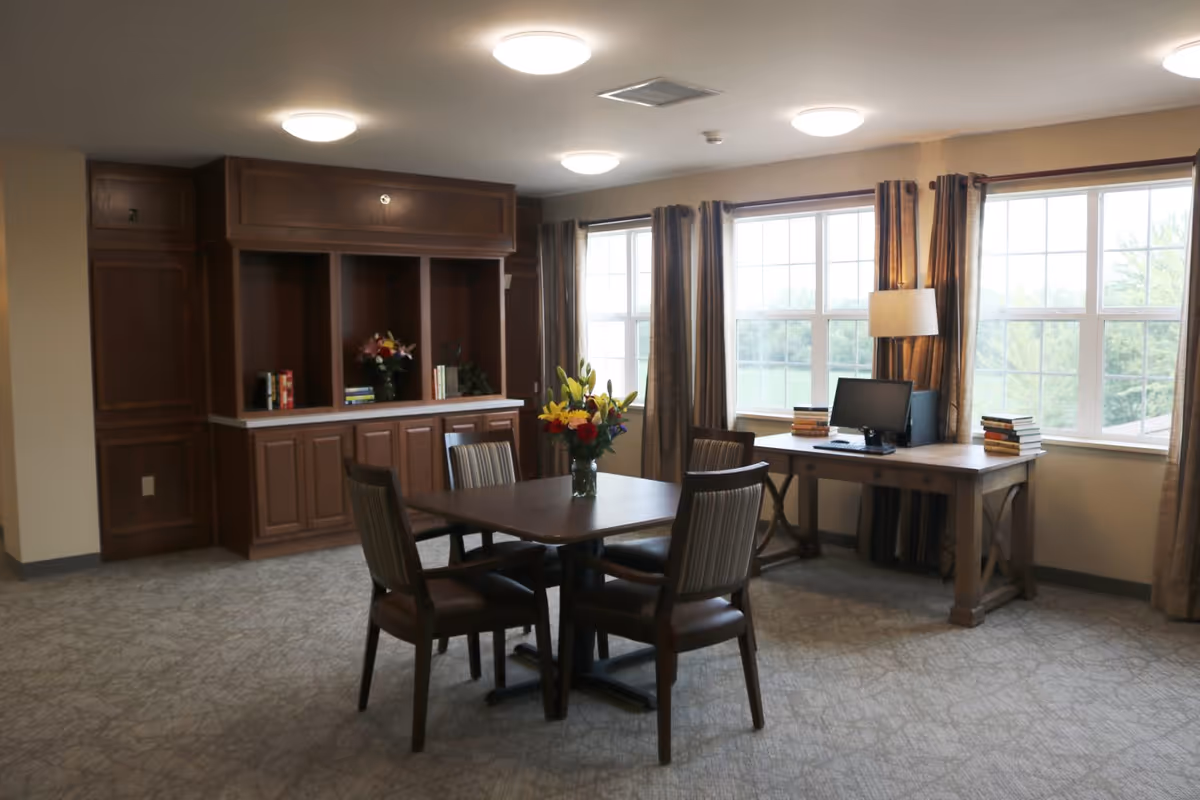 A well-lit common room with a square wooden table surrounded by four chairs, a vase with colorful flowers on the table, large windows with curtains letting in natural light, a wooden desk with a computer and stacks of books, and built-in wooden cabinets along the wall.